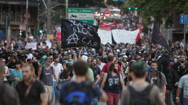 Eine große Menschenmenge demonstriert mit einer „Black Bloc“-Flagge in einer städtischen Straße.