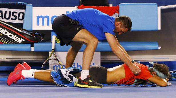 Ein Tennisspieler liegt auf dem Boden, während ein Trainer seine Muskeln behandelt.