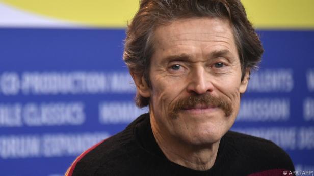 Porträt von Willem Dafoe mit Schnurrbart vor blauem Hintergrund.