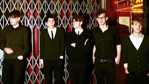 Fünf junge Männer posieren vor einem Gitter und einem beleuchteten Eingang.