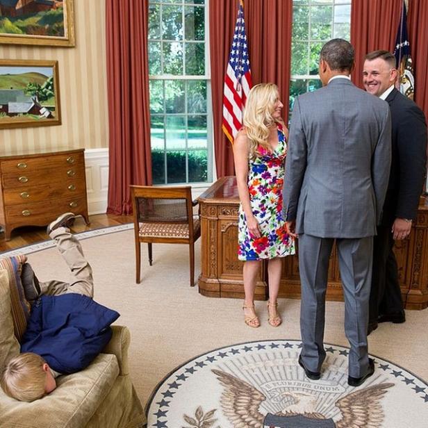 Ein Junge macht einen Handstand auf einem Sofa, während Barack Obama sich mit zwei anderen Personen unterhält.