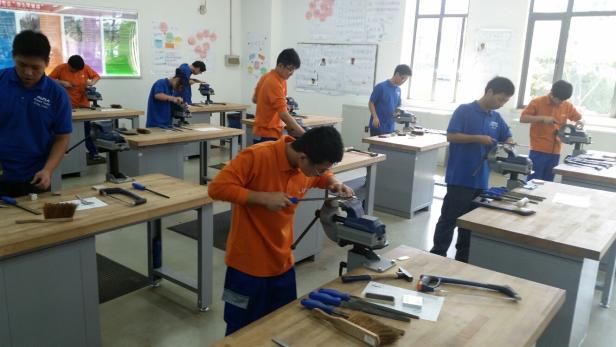 Schüler arbeiten in einer Werkstatt an Werkbänken mit Schraubstöcken und Werkzeugen.
