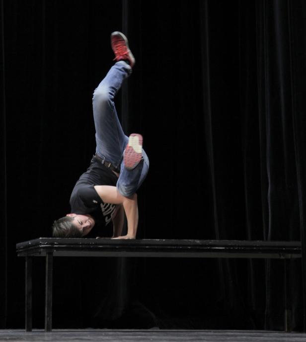Eine Frau macht einen Handstand auf einem Tisch vor einem schwarzen Vorhang.