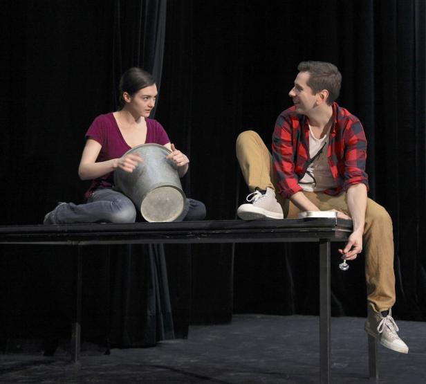 Eine Frau mit einem Eimer und ein Mann sitzen auf einem Tisch vor einem schwarzen Vorhang.