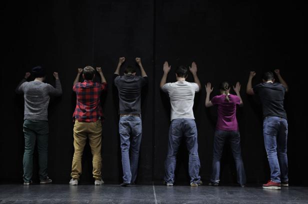 Eine Gruppe von sechs Personen steht mit erhobenen Händen vor einer schwarzen Wand.