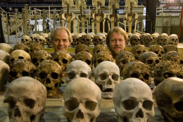 Zwei Männer stehen hinter einer großen Anzahl von Schädeln und Skeletten.
