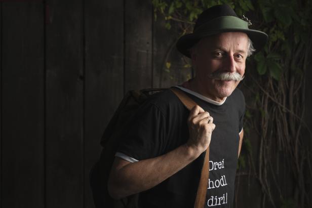 Ein Mann mit Hut, Schnurrbart und Rucksack vor einer Holzwand.