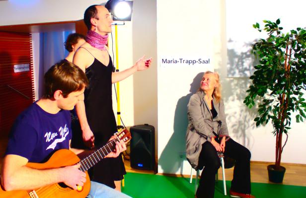 Ein Mann spielt Gitarre, während eine Person im Kleid spricht und eine Frau im Maria-Trapp-Saal zuhört.