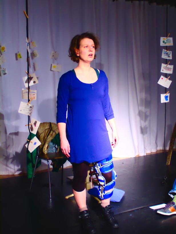 Eine Frau mit einer Knieorthese steht auf einer Bühne vor einem weißen Vorhang.