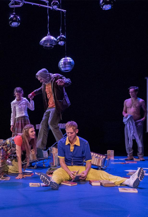 Eine Theaterszene mit mehreren Schauspielern auf einer Bühne voller Bücher.