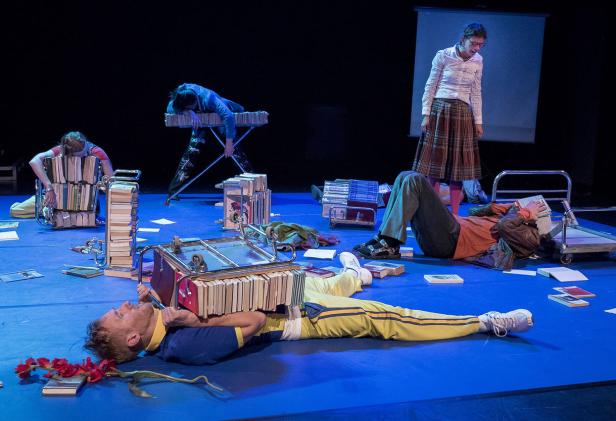 Mehrere Schauspieler liegen auf einer Bühne, umgeben von Büchern und Bücherwagen.