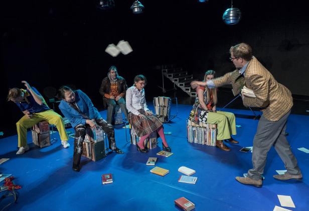 Eine Theaterszene mit mehreren Personen, die auf Bücherstapeln sitzen und mit Büchern interagieren.