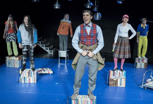 Mehrere Schauspieler stehen auf einem Bühne, die mit Büchern und einem blauen Boden ausgelegt ist.