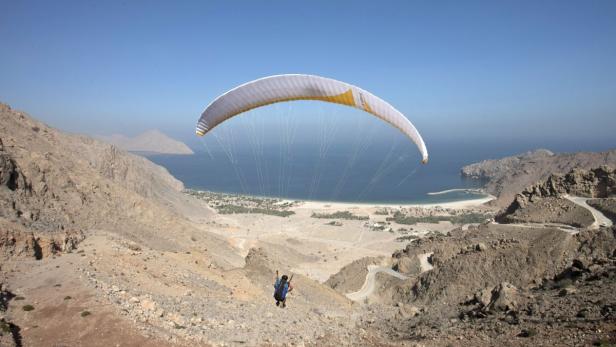 Ein Gleitschirmflieger startet von einem felsigen Hang mit Blick auf eine Küstenstadt.