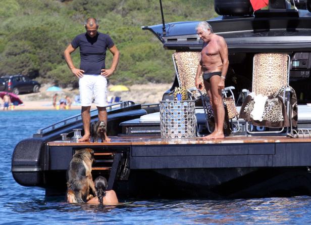 Zwei Männer und zwei Hunde auf einer Yacht, während eine Frau im Wasser steht.