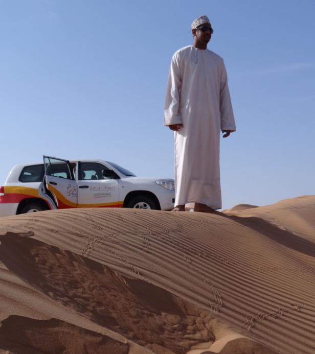 Ein Mann in traditioneller Kleidung steht in der Wüste neben einem Geländewagen von Zahara Tours.