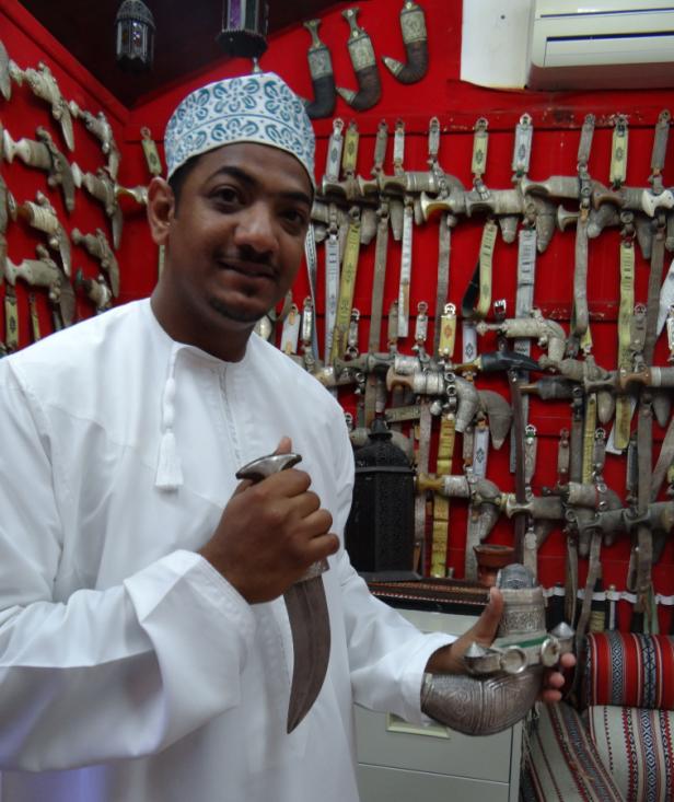 Ein Mann in traditioneller Kleidung präsentiert einen Krummdolch vor einer Wand mit weiteren Dolchen.