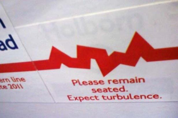 Ein Schild mit der Aufschrift „Please remain seated. Expect turbulence.“ und einer roten Zickzacklinie.