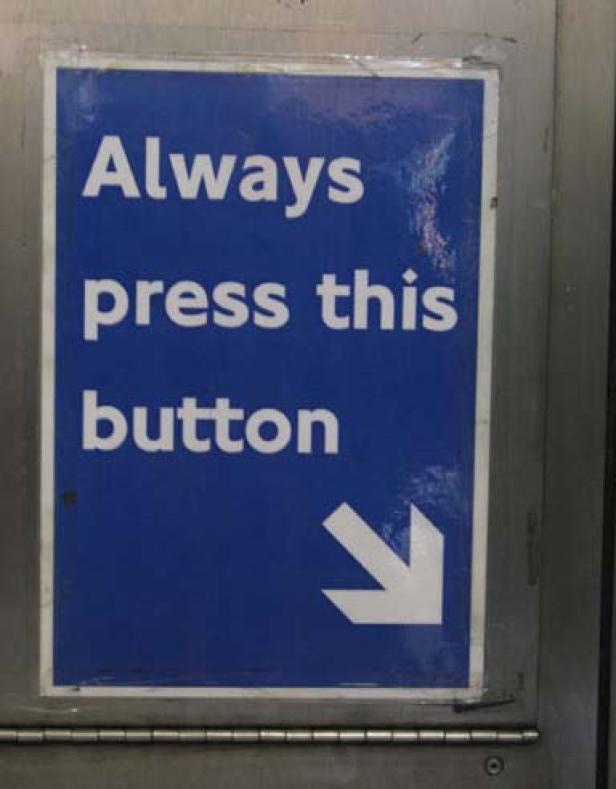 Ein blaues Schild fordert mit einem Pfeil auf, einen Knopf zu drücken.