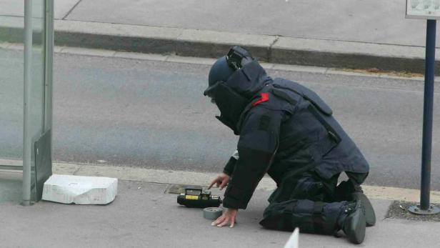 Ein Bombenentschärfer in Schutzkleidung untersucht einen verdächtigen Gegenstand auf dem Bürgersteig.