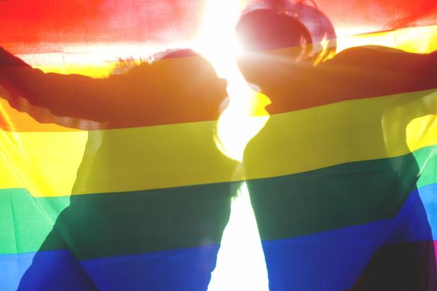 Zwei Silhouetten halten eine Regenbogenflagge im Gegenlicht.