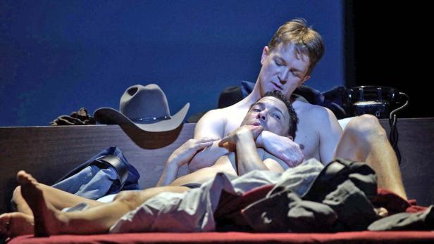 Zwei Männer liegen zusammen in einem Bett, ein Cowboyhut ist im Hintergrund zu sehen.