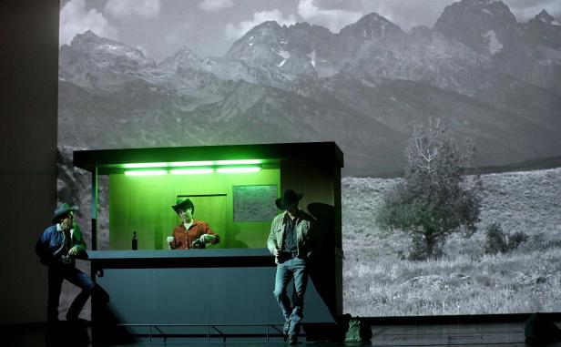 Eine Bühnenszene mit drei Personen in Cowboyhüten vor einer Bergkulisse.