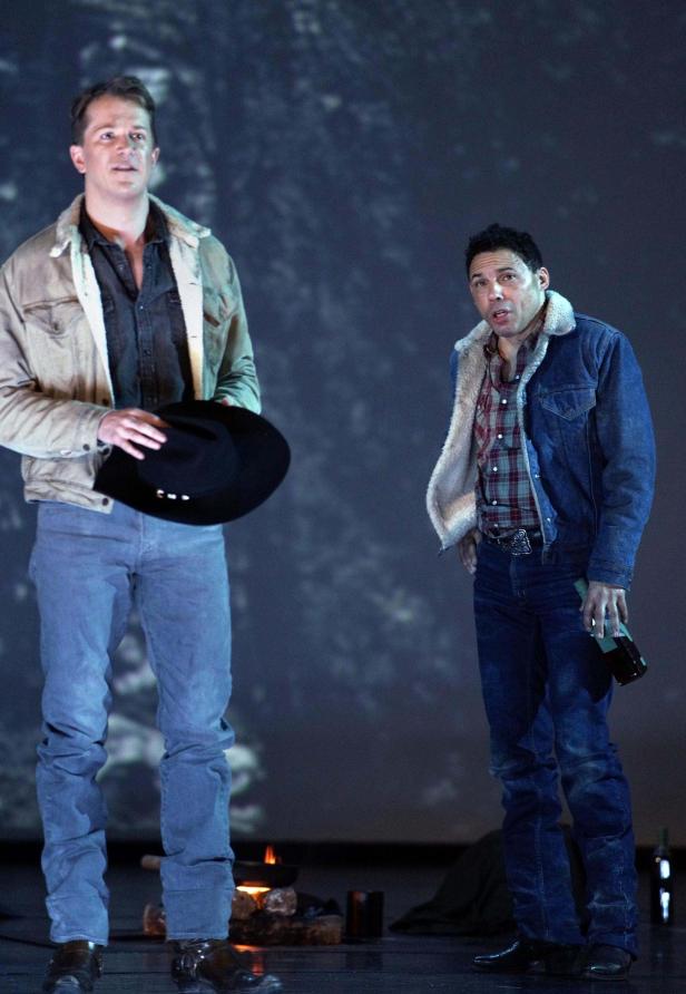 Zwei Männer in Jeansjacken stehen vor einem Lagerfeuer.