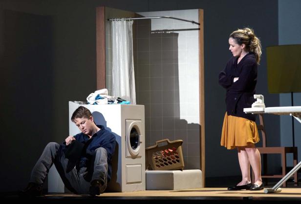 Auf einer Bühne sitzt ein Mann neben einer Waschmaschine, während eine Frau mit verschränkten Armen steht.