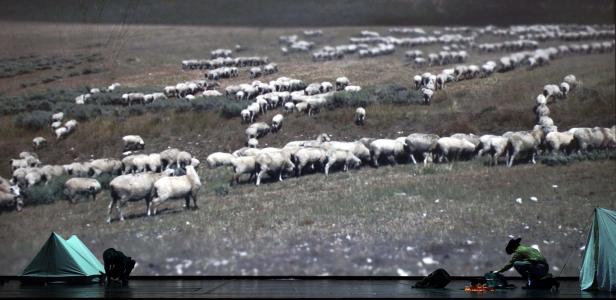 Auf einer Bühne zündet eine Person neben Zelten ein Lagerfeuer vor einer Projektion einer Schafherde an.