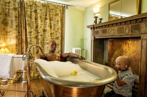Eine Schaufensterpuppe entspannt in einer mit Schaum gefüllten Badewanne, während eine andere Puppe zusieht.