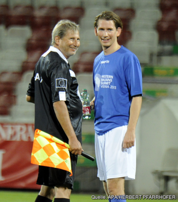 Ein Schiedsrichter mit Fahne und Sebastian Kurz in Sportkleidung stehen auf einem Fußballfeld.