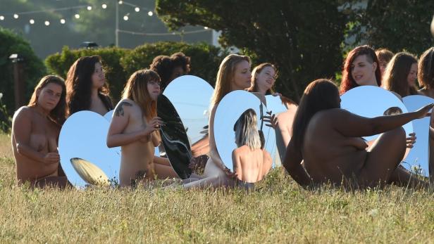 Eine Gruppe unbekleideter Frauen sitzt mit runden Spiegeln auf einer Wiese.