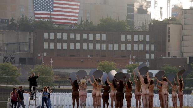 Eine Gruppe nackter Frauen hält graue Scheiben vor einer Industrie- und Stadtkulisse mit US-amerikanischer Flagge.