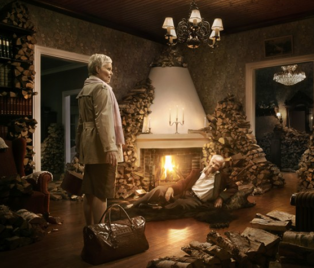 Eine Frau steht in einem mit Holzscheiten dekorierten Raum, während ein Mann am Kamin liegt.