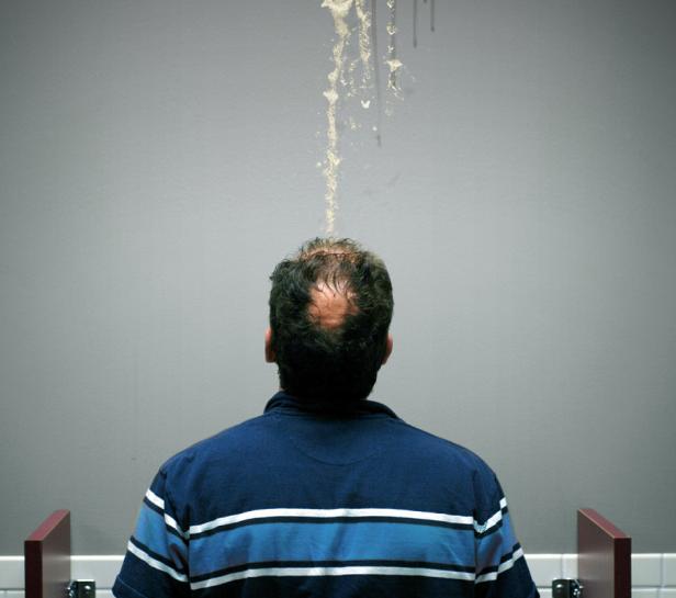 Ein Mann steht in einem Waschraum, während Flüssigkeit von der Decke auf seinen Kopf tropft.