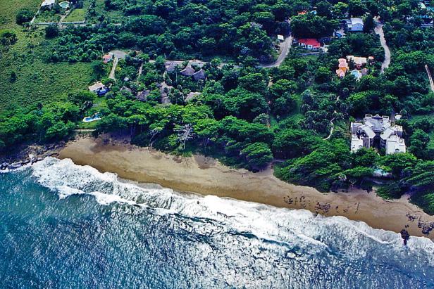 Luftaufnahme einer Küstenlinie mit Strand, dichter Vegetation und einigen Häusern.