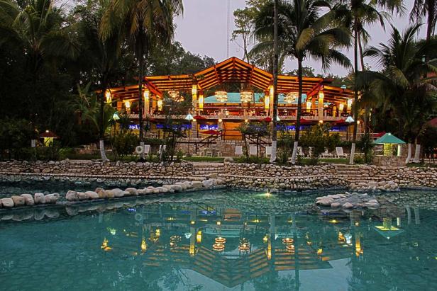 Ein beleuchtetes Gebäude mit Restaurant am Ufer eines Sees, umgeben von Palmen.