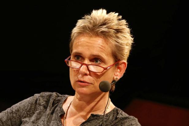 Eine Frau mit blonden, stacheligen Haaren und roter Brille spricht in ein Mikrofon.