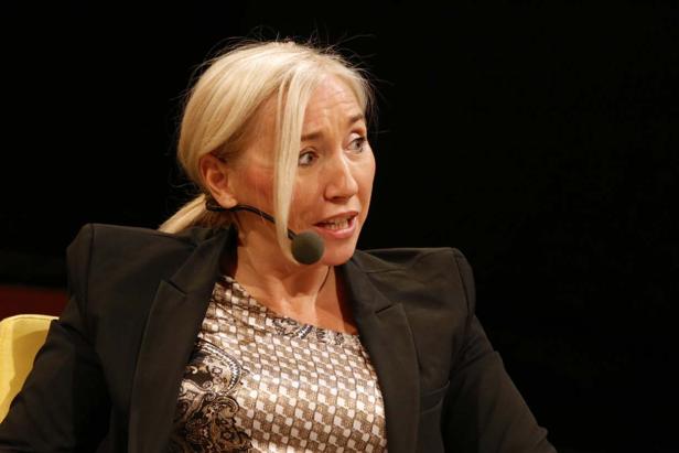 Eine blonde Frau mit Mikrofon spricht vor einem dunklen Hintergrund.