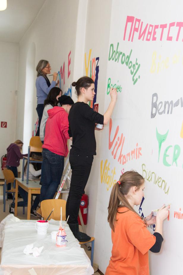 Mehrere Personen bemalen eine Wand mit dem Wort „Willkommen“ in verschiedenen Sprachen.