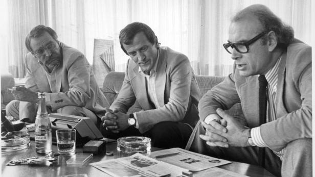 Drei Männer sitzen an einem Tisch mit Getränken und Papieren, vermutlich in einer Besprechung.