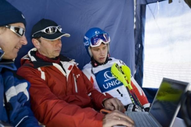 Drei Personen, darunter ein Skirennläufer, sitzen vor einem Laptop in einem Zelt.