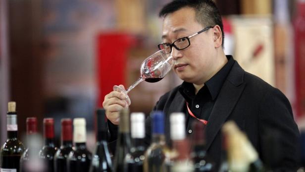 Ein Mann mit Brille riecht an einem Glas Rotwein vor einer Reihe von Weinflaschen.