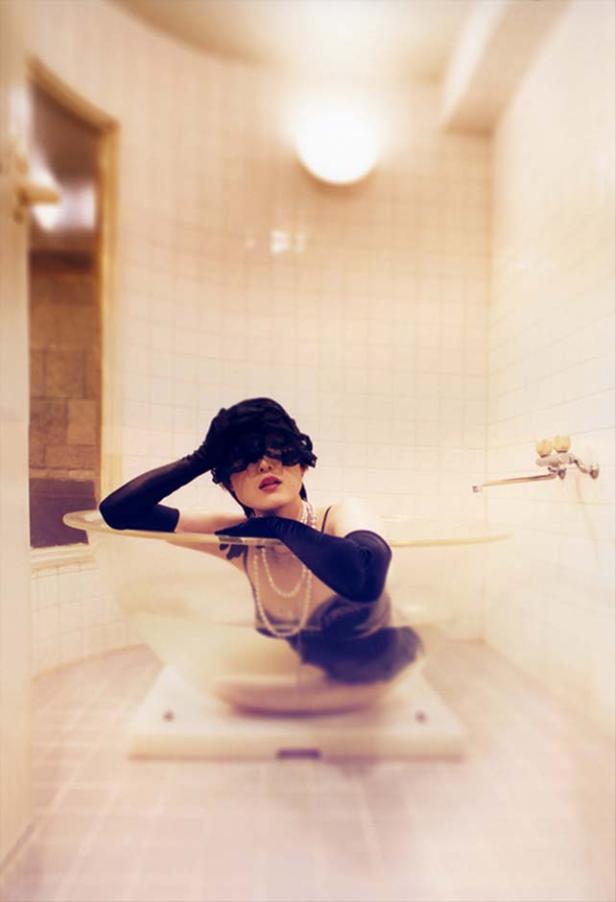 Eine Frau mit Hut, Handschuhen und Perlenkette entspannt in einer Badewanne.