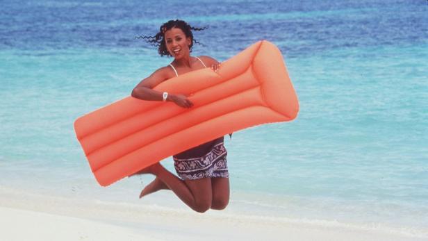 Eine Frau springt am Strand mit einer orangefarbenen Luftmatratze in der Hand.