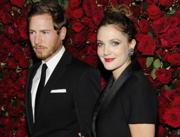Drew Barrymore und ein Mann in Anzügen posieren vor einer Wand aus roten Rosen.