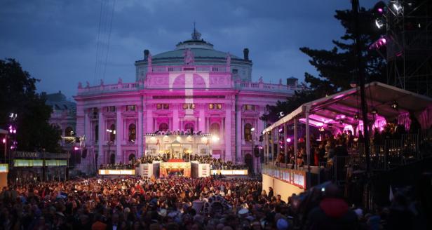 Das Wiener Opernhaus ist während einer Veranstaltung mit rosa Licht beleuchtet; eine große Menschenmenge ist davor versammelt.