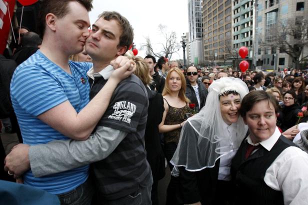 Zwei Männer umarmen sich auf einer Kundgebung, im Hintergrund weitere Menschen und Ballons.