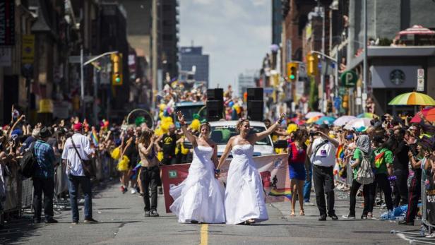 Zwei Frauen in weißen Kleidern führen eine Parade an, umgeben von Zuschauern und bunten Dekorationen.